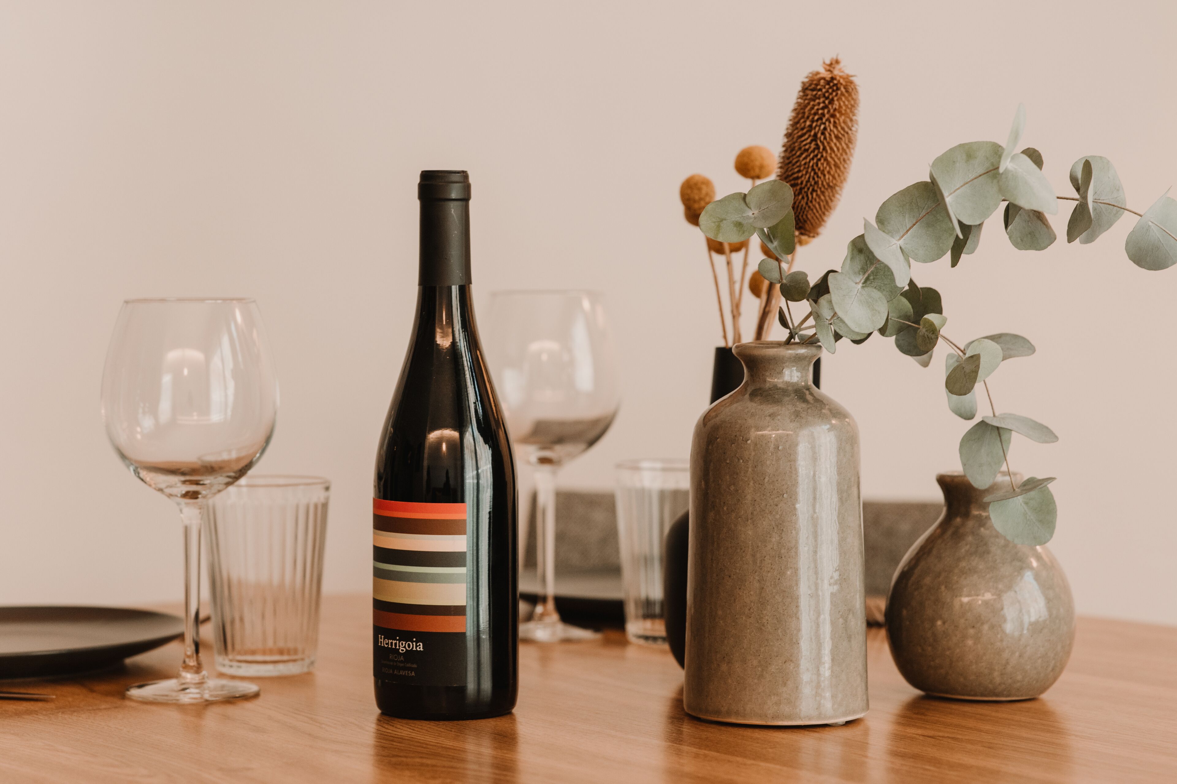 A wine bottle, two wine glasses, and a vase with eucalyptus leaves on a wooden table.