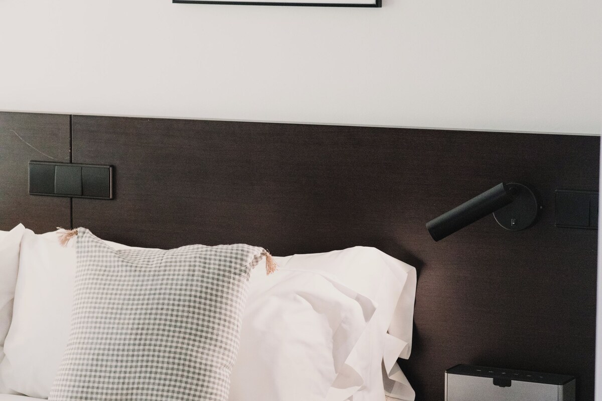 Moderni hotellihuone, jossa on siististi pedattu sänky, yöpöytä lampun kanssa ja seinällä oleva kehystetty abstrakti taideteos.