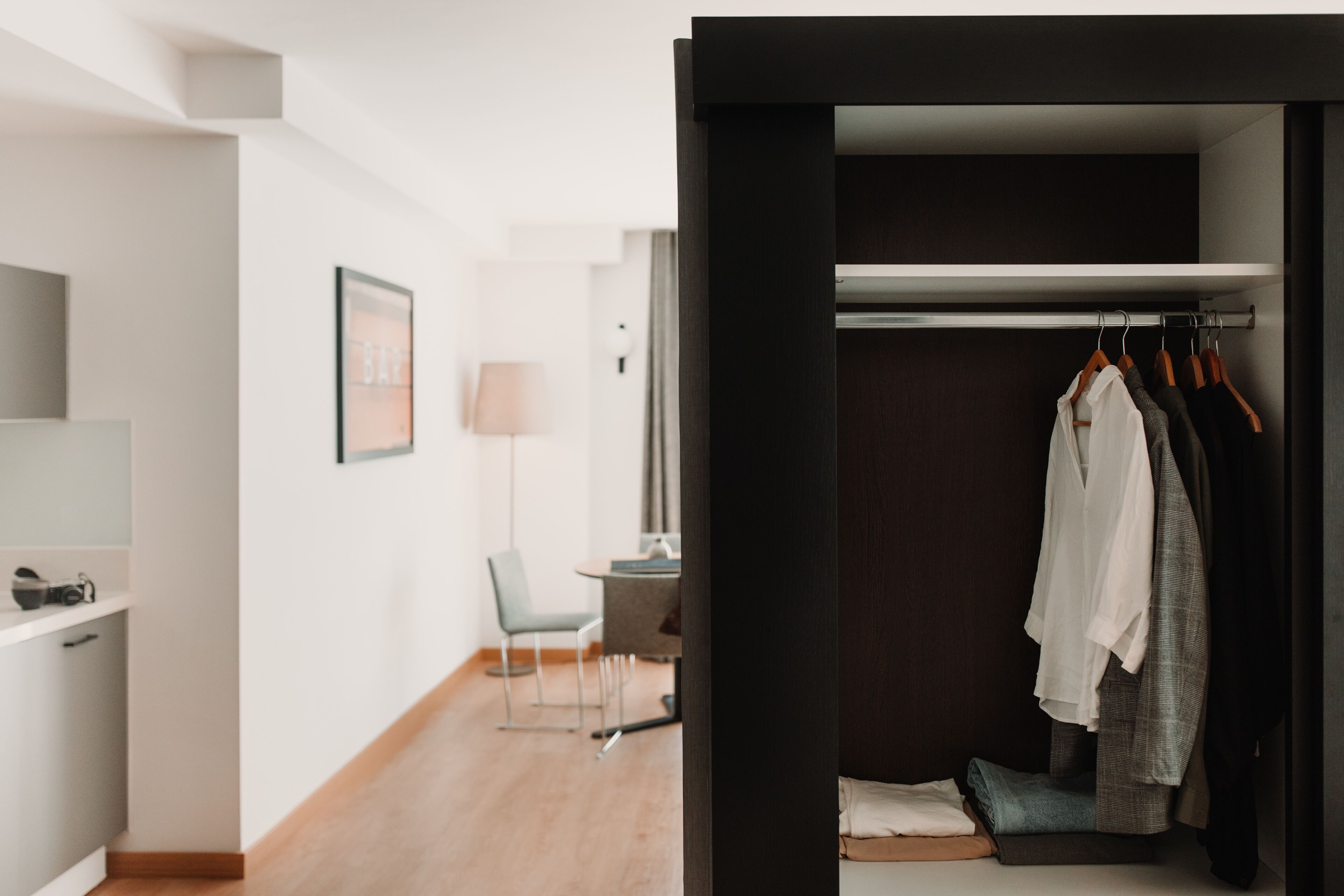 Habitación de hotel moderna con piso de madera, un escritorio con lámpara, una silla y un armario con ropa colgada.