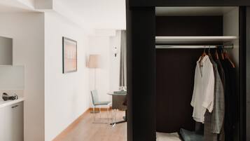 Habitación de hotel moderna con piso de madera, un escritorio con lámpara, una silla y un armario con ropa colgada.