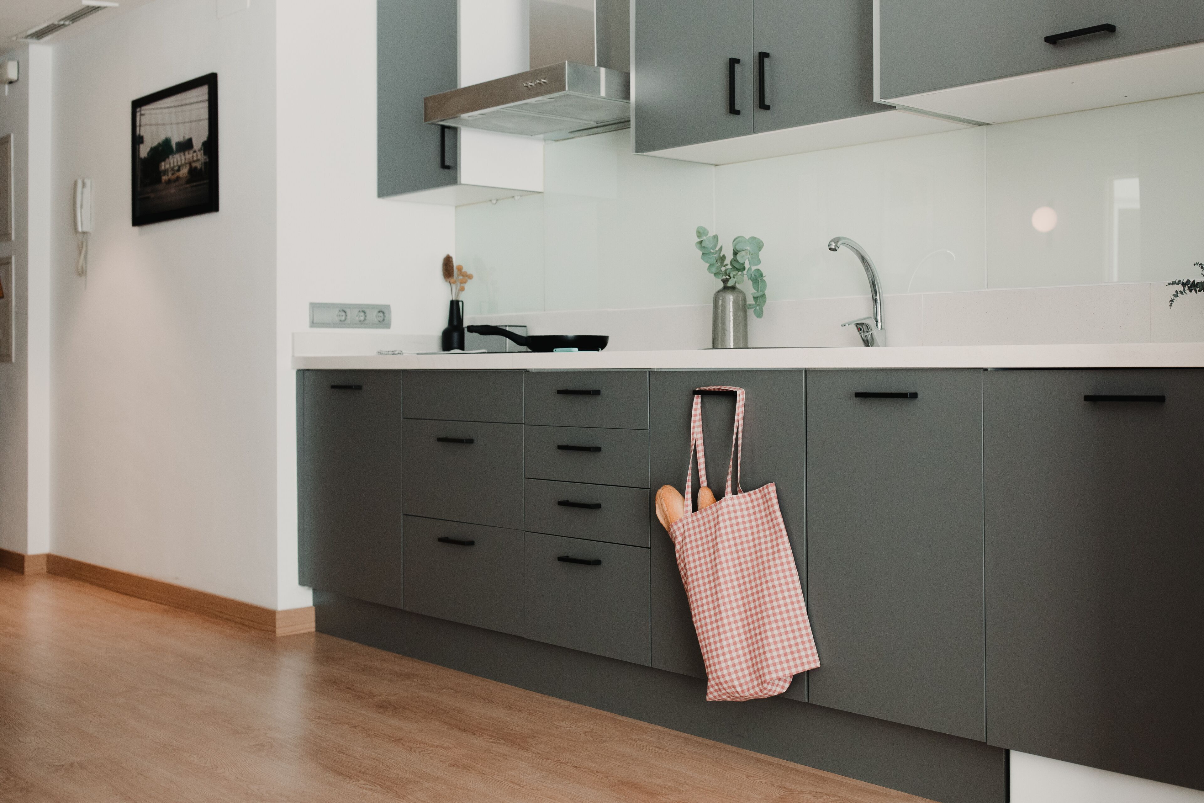 A modern kitchen with grey cabinets, a stainless steel range hood, a white countertop, and a checkered bag hanging on the cabinet door.