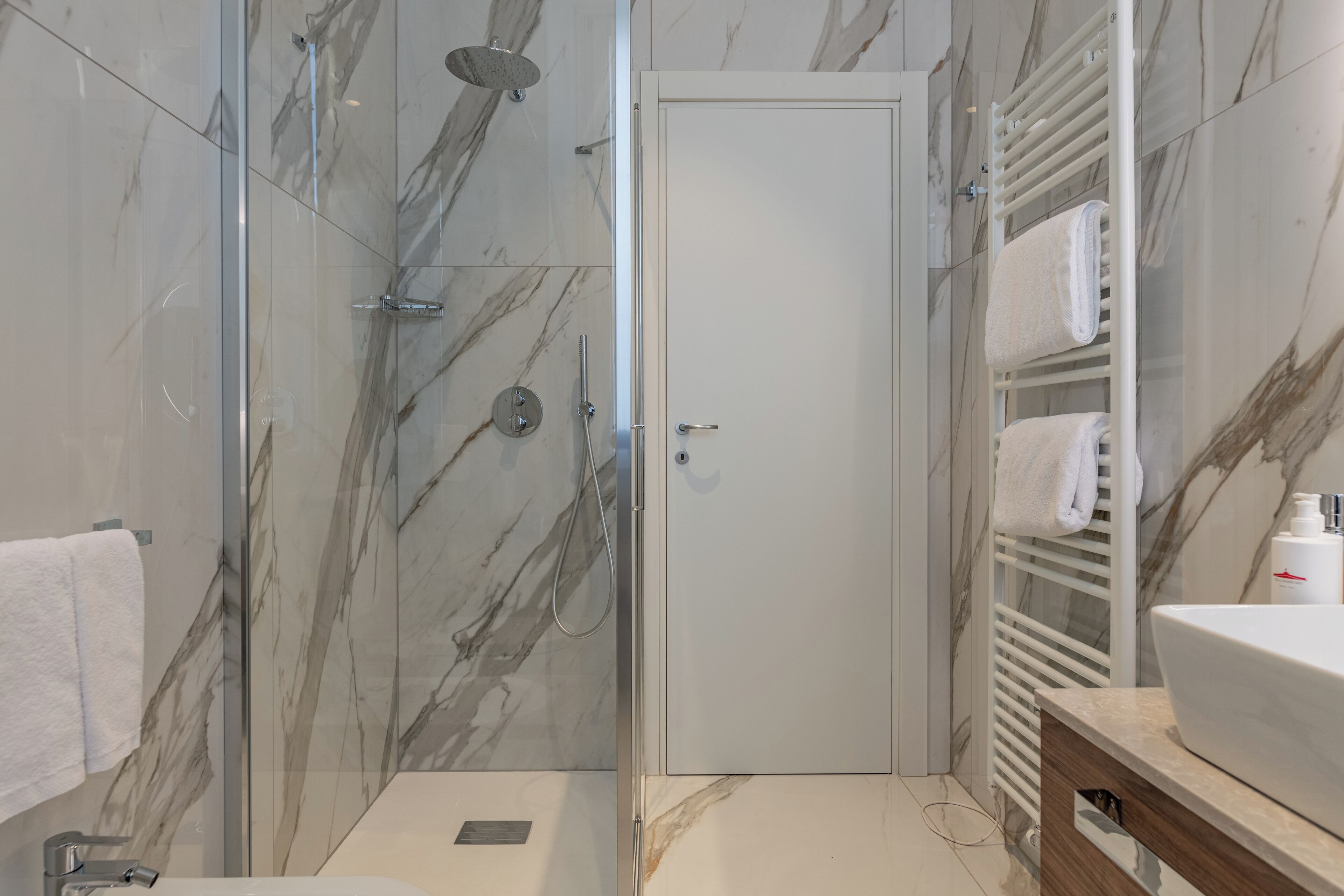 Un baño moderno con ducha envuelta en vidrio, paredes de mármol, una puerta blanca y un lavabo con toallero.