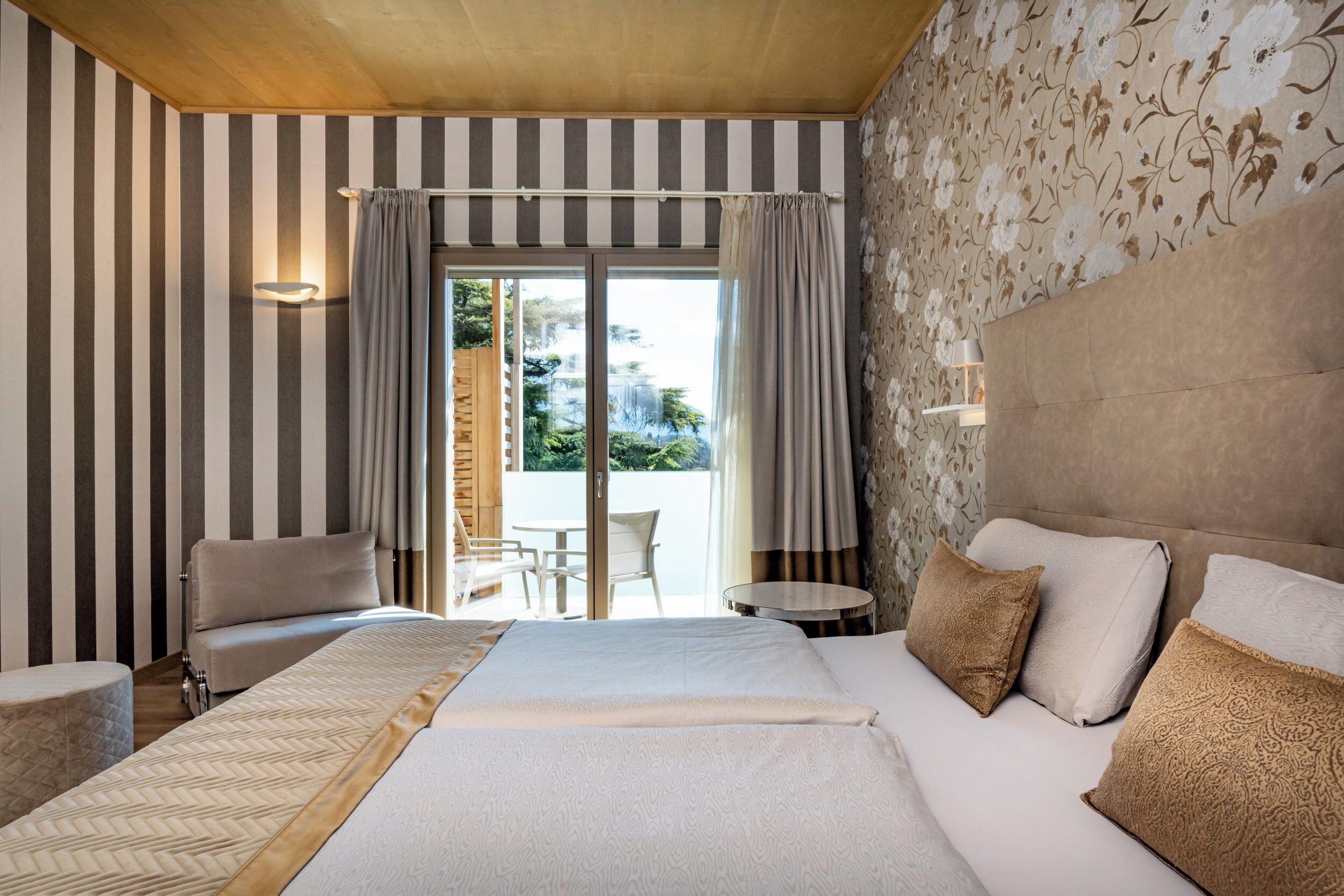 A hotel room with a large bed, a sofa, a small table, and a view of the outdoors through a sliding door.