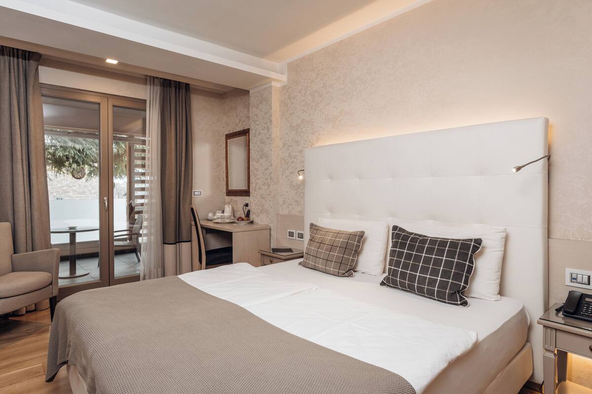 Ein Hotelzimmer mit einem großen Bett, einem Schreibtisch mit Stuhl, einem kleinen Tisch und Blick durch eine Glastür.