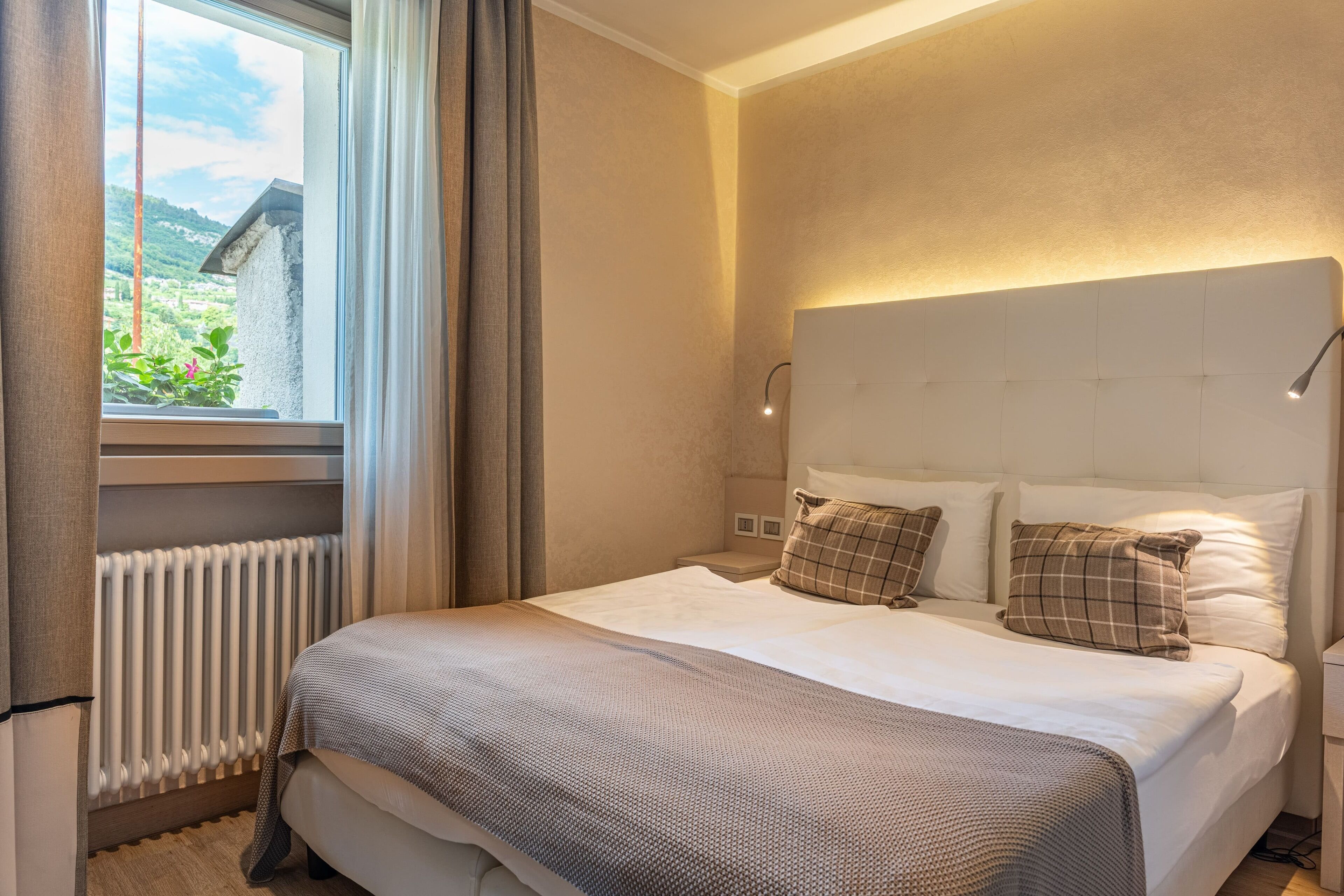 Un dormitorio con cama, ventana con cortinas, radiador y una lámpara fijada en la pared.
