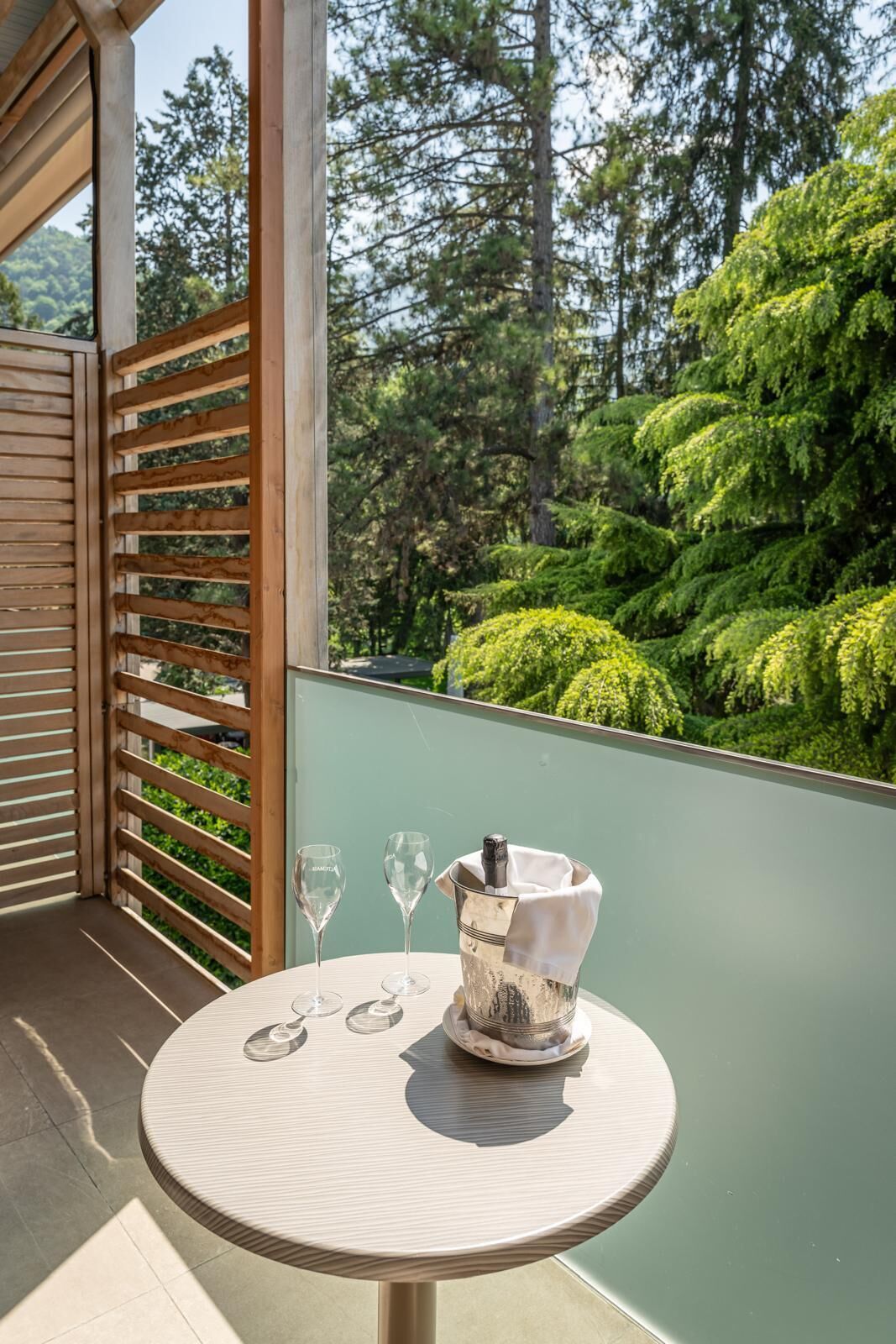 A round table with a bottle of champagne and two glasses on a balcony with a view of a forest.