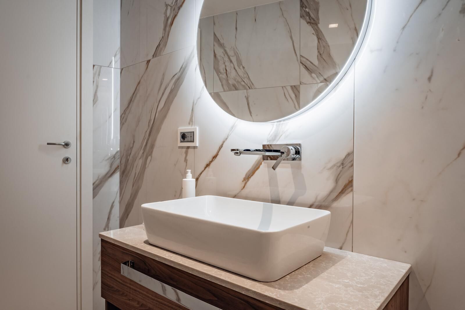 A bathroom with a white vessel sink, a wooden vanity, a marble wall, and a round mirror.