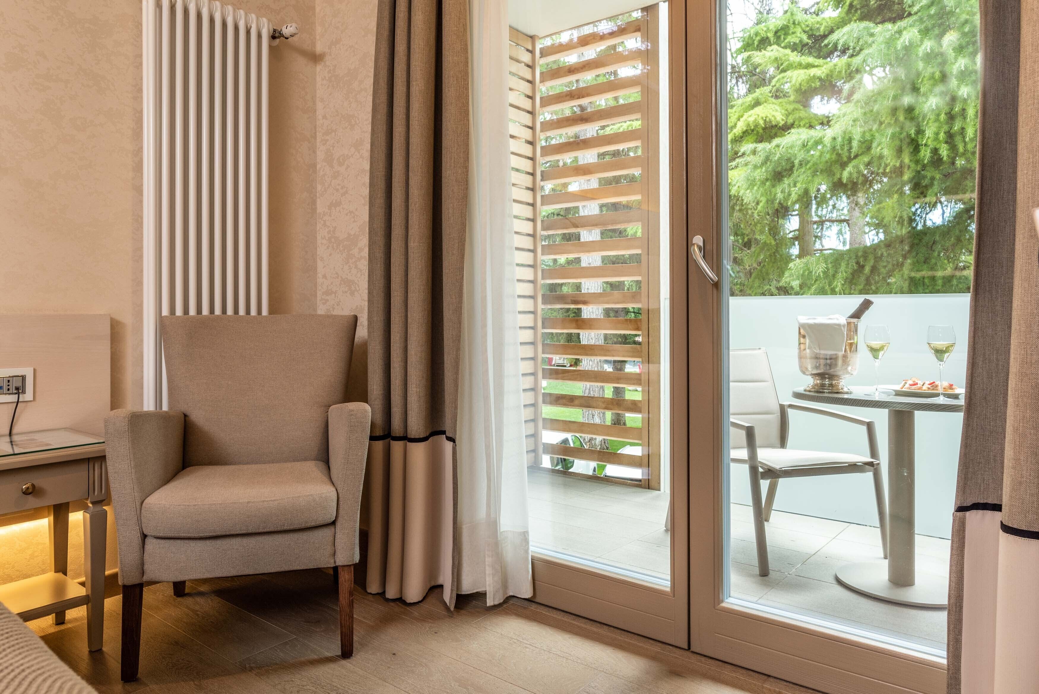 Una stanza con una porta scorrevole in vetro che dà su un balcone, una poltrona beige, un tavolino con piano in vetro e vista sul verde circostante.
