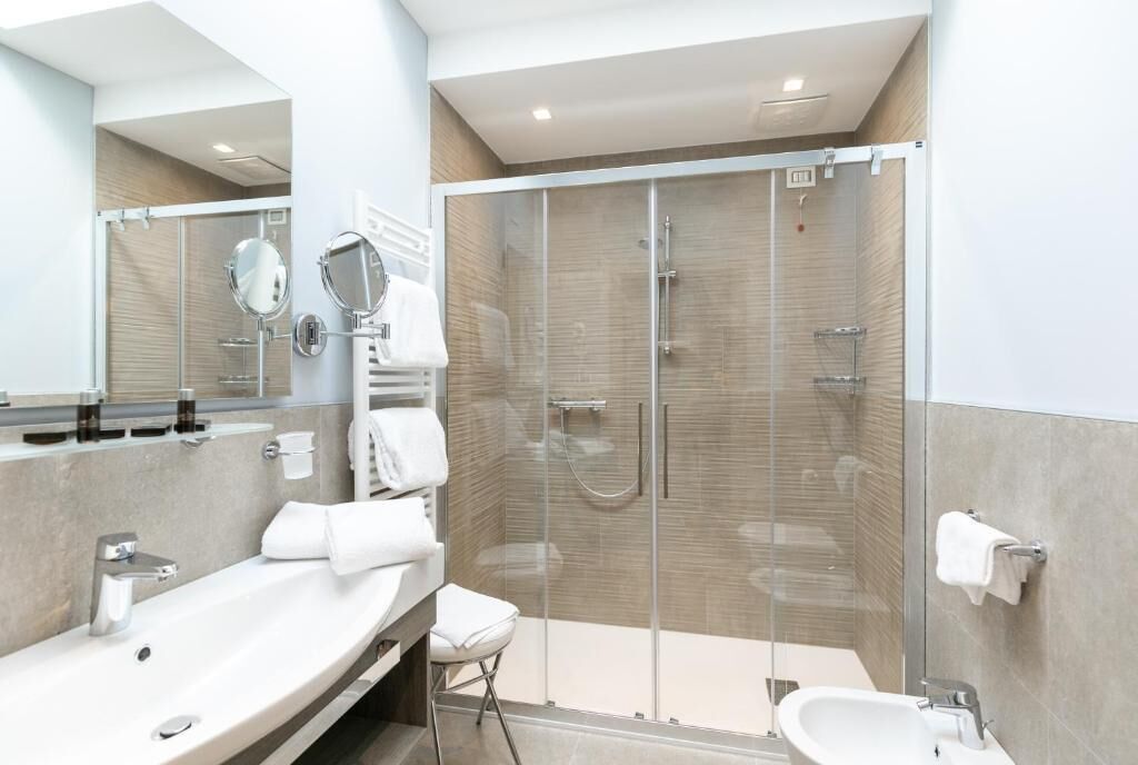 A modern bathroom with a large mirror, two sinks, a shower enclosure, and a small stool.