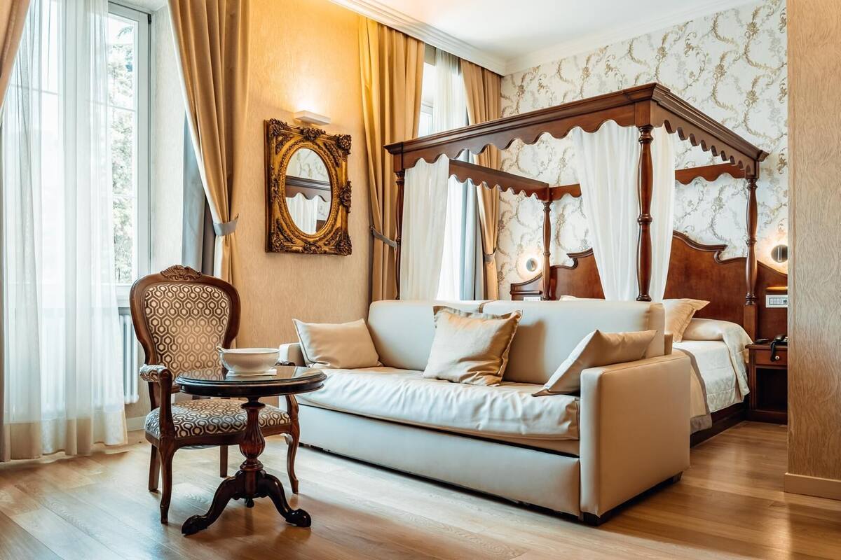 Ein Bett mit Himmelbett und Holzfußteil, eine Couch, ein Sessel und ein kleiner Tisch in einem gut beleuchteten Raum mit gemustertem Wandpapier und Vorhängen.