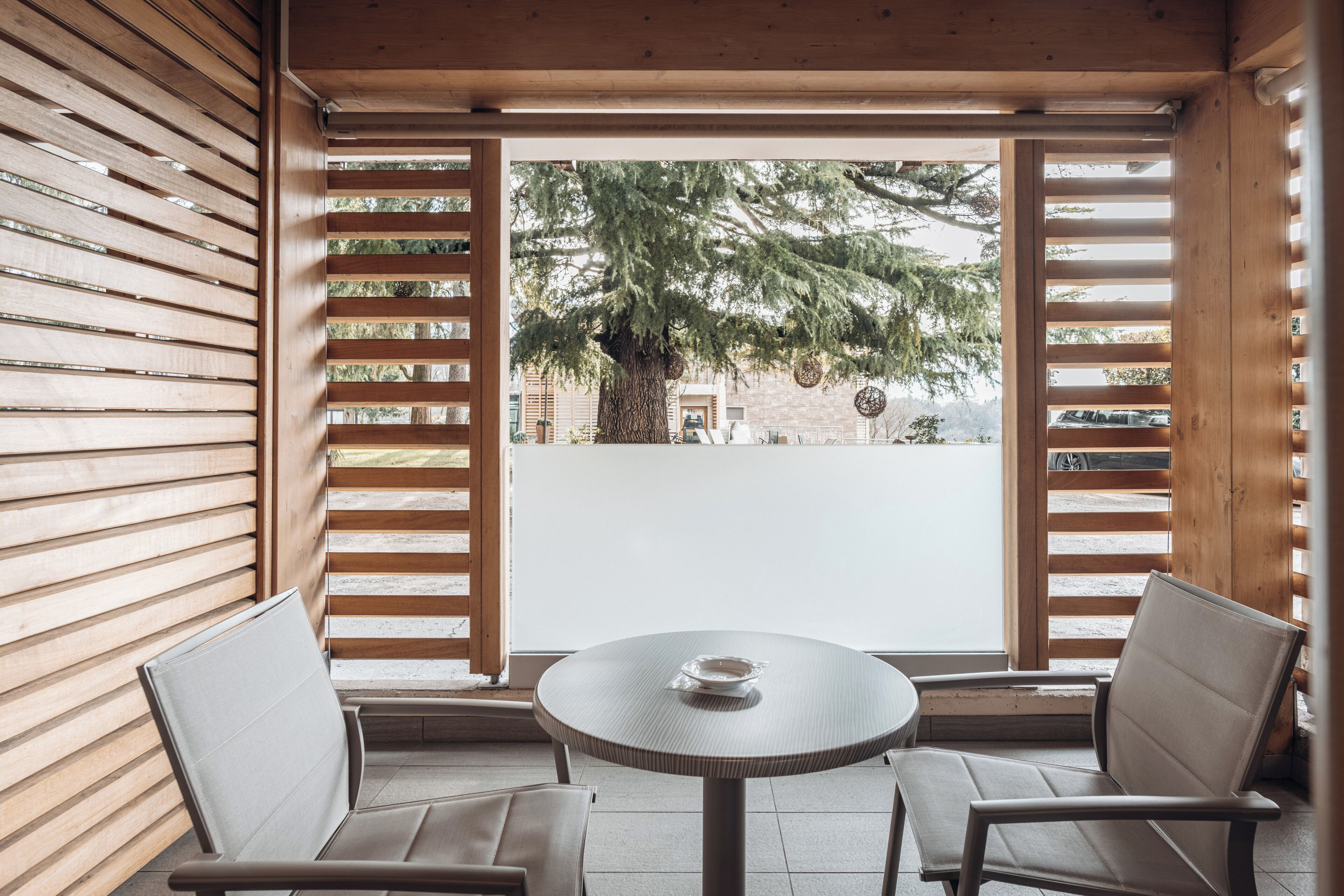 Un espacio al aire libre acogedor con una mesa redonda y dos sillas, listones de madera en las paredes, y vistas a un árbol y un edificio.