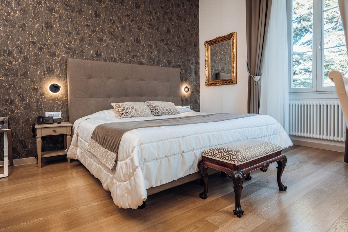 Ein Schlafzimmer mit einem großen Bett, Nachttischen, einer Bank, einem Spiegel und einem Fenster mit Vorhängen.
