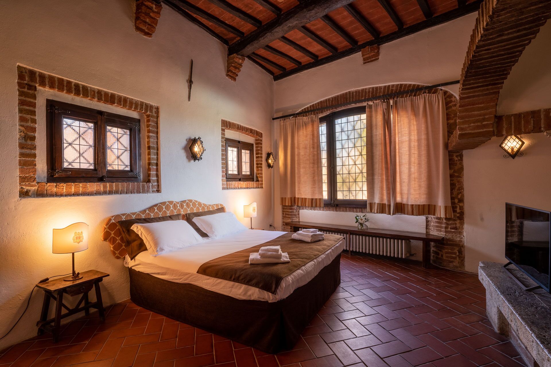 Una habitación con una cama, una mesita de noche con lámpara, una chimenea y un ventanal con cortinas.