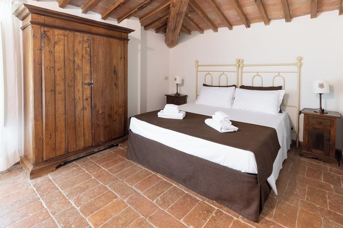 A bedroom with a wooden wardrobe, a bed with white and brown linens, and a nightstand with a lamp.