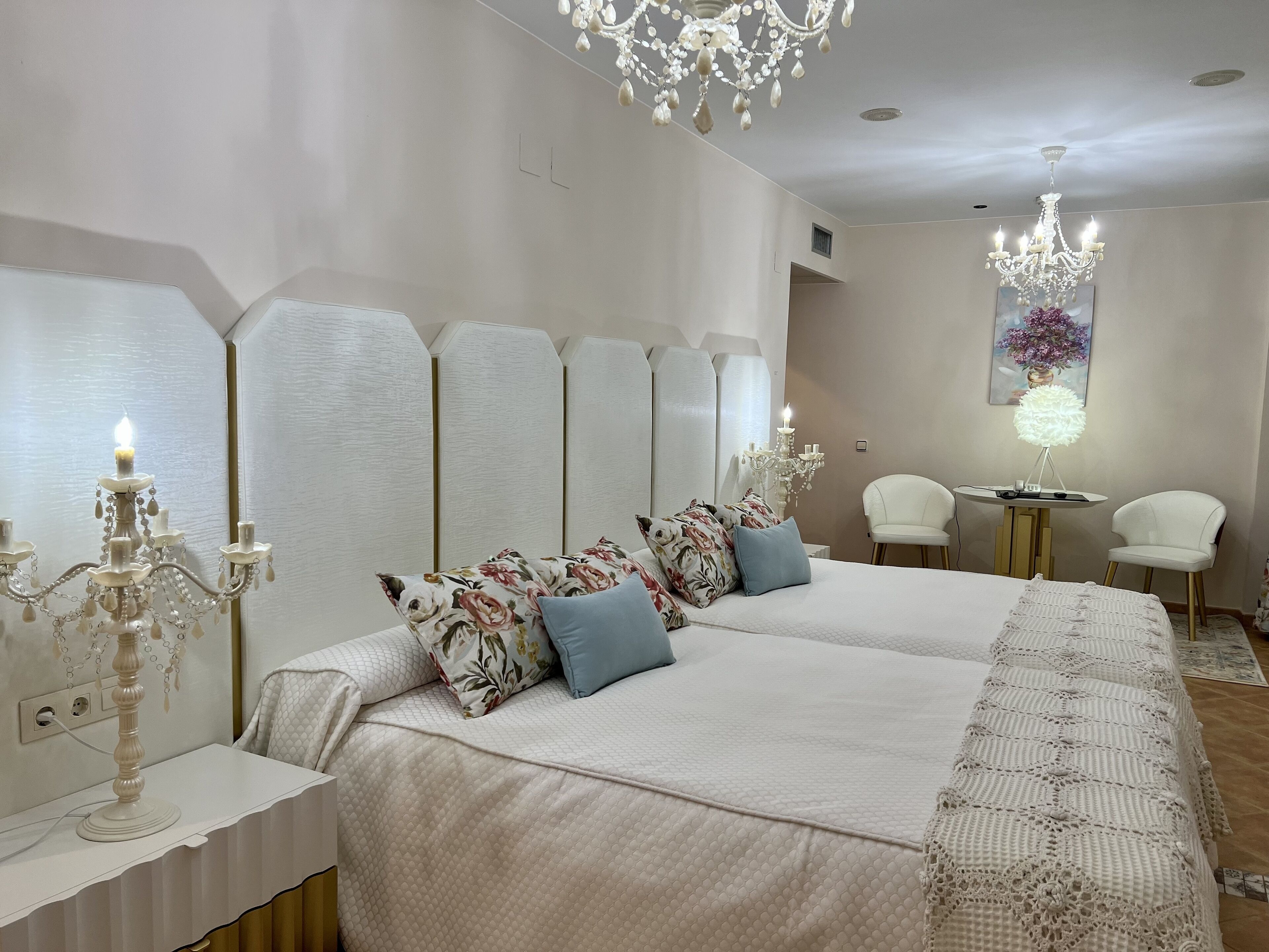 A hotel room with a large bed, two bedside tables, a chandelier, a small table with a lamp, and two chairs.