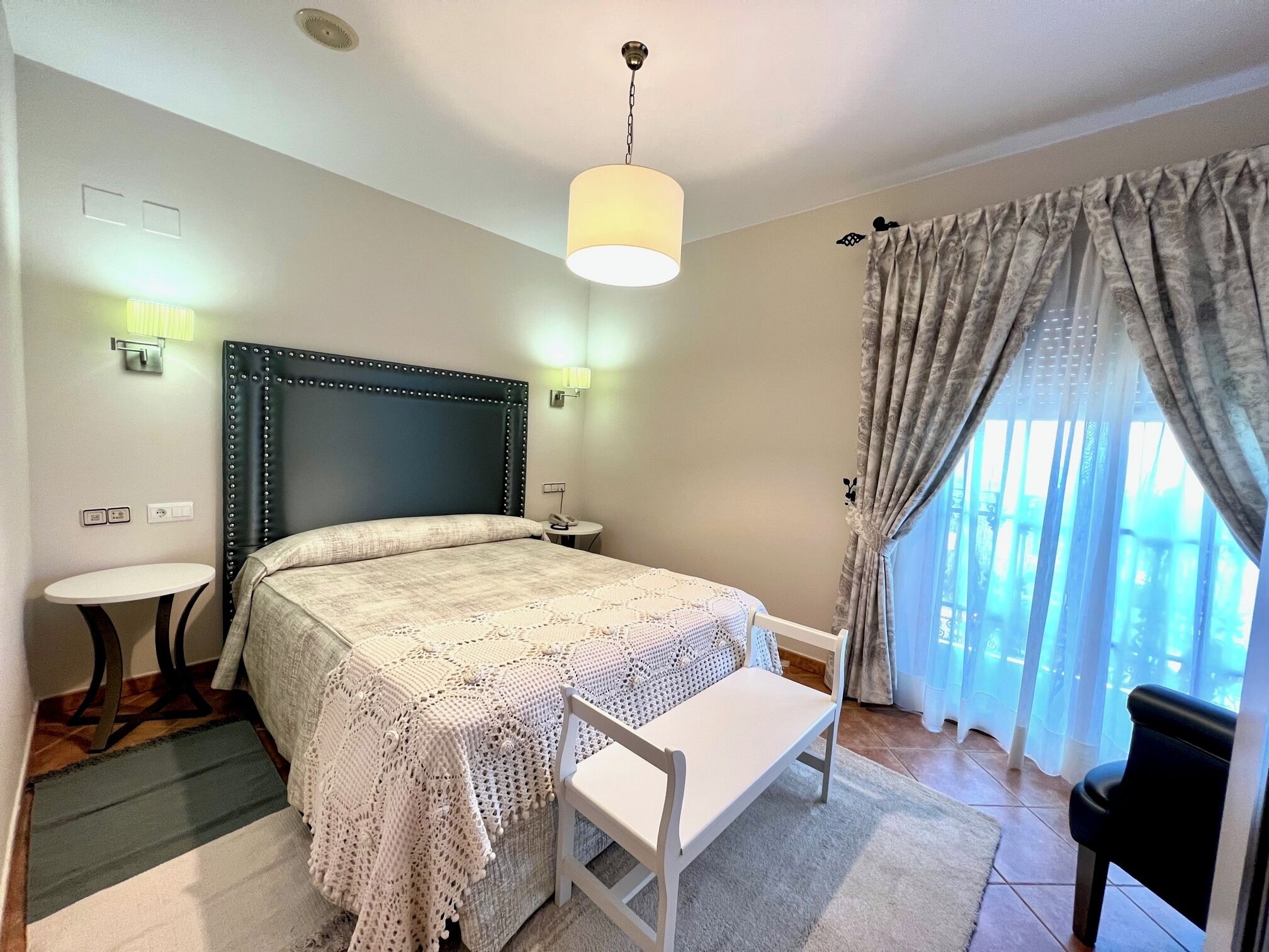 Habitació d'hotel amb llit, taules de nit, escriptori, cadira, làmpada, cortines i finestra.