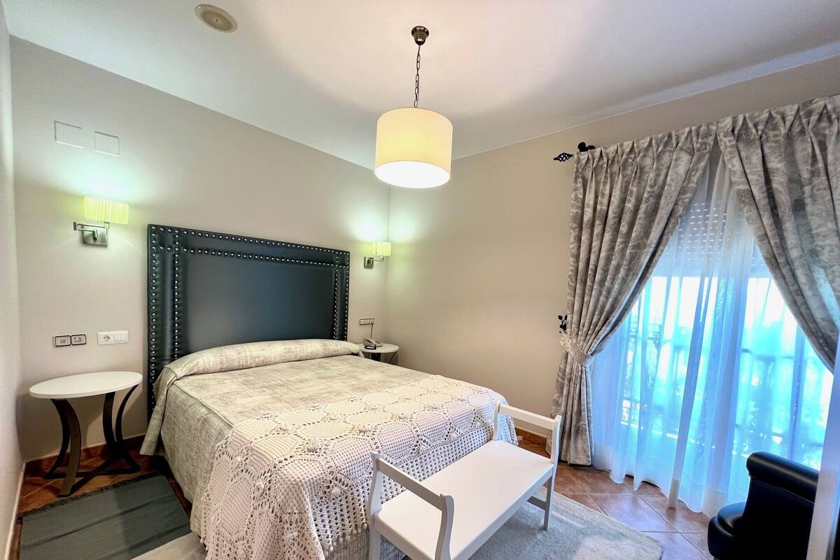 A hotel room with a bed, bedside tables, a desk, a chair, a lamp, curtains, and a window.