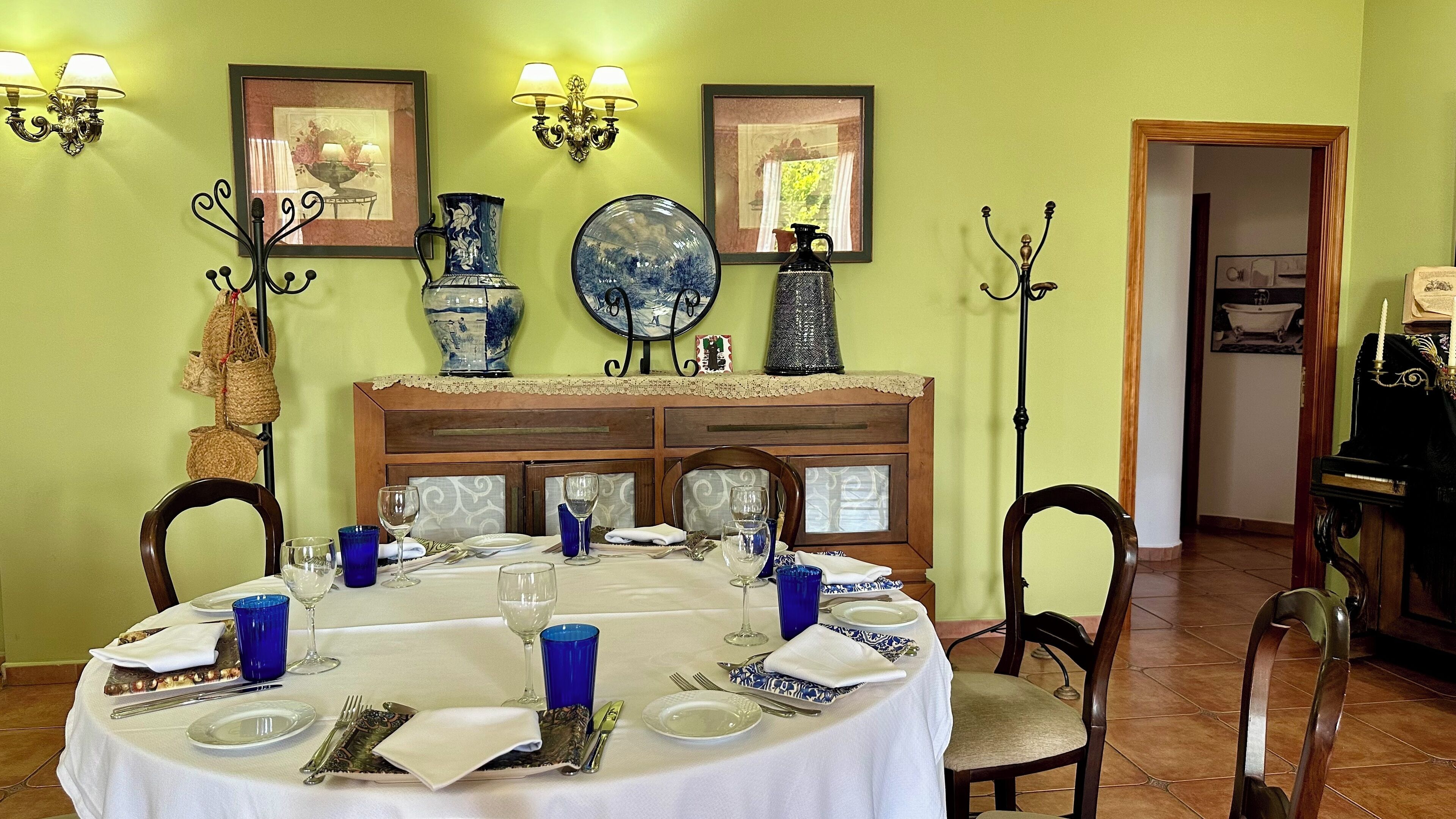 A dining room with a round table set for four, surrounded by chairs. There are wall-mounted light fixtures and framed pictures above the table.