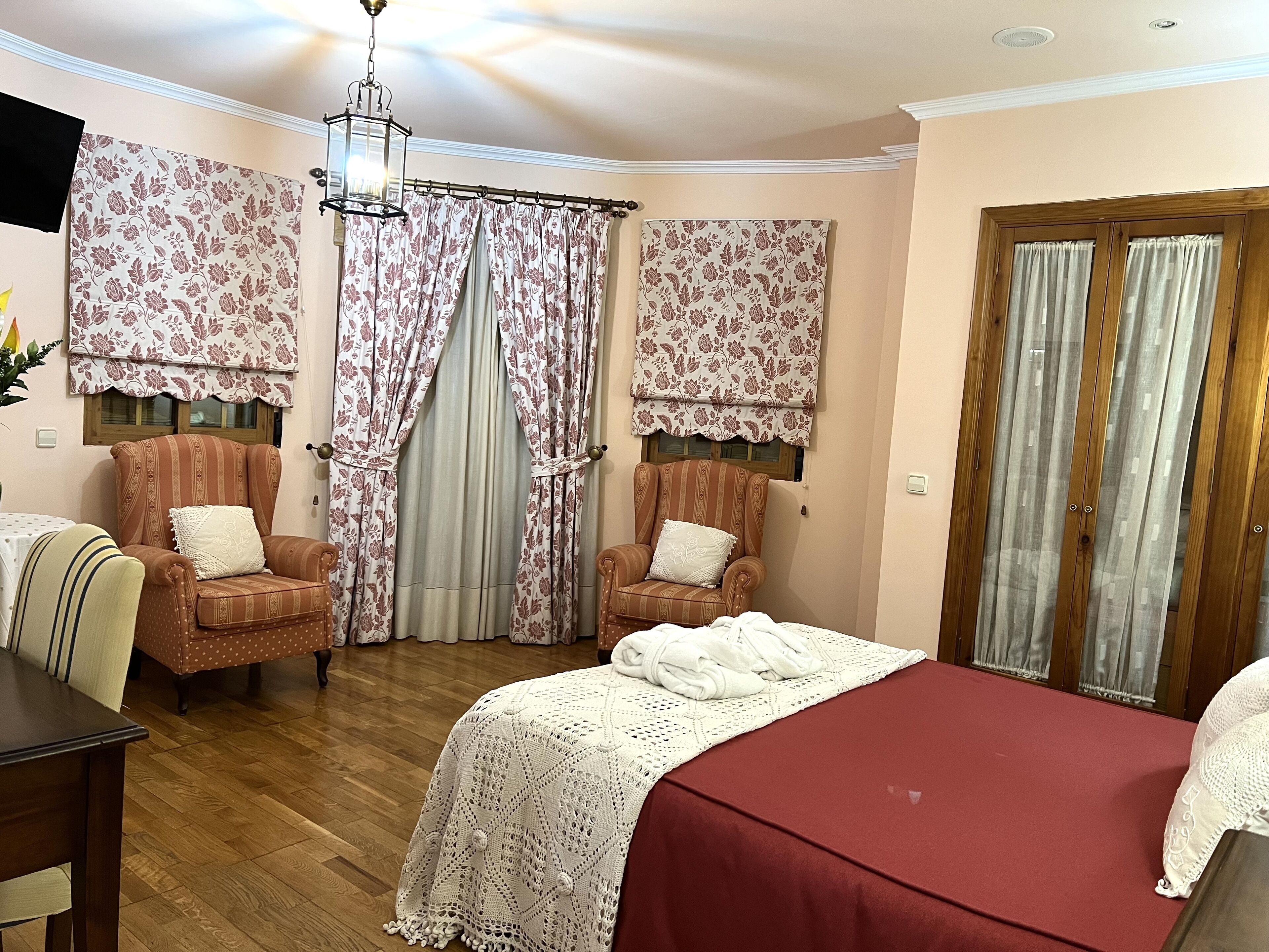 A bedroom with two armchairs, a bed with a red bedspread, floral curtains, and a wooden door.