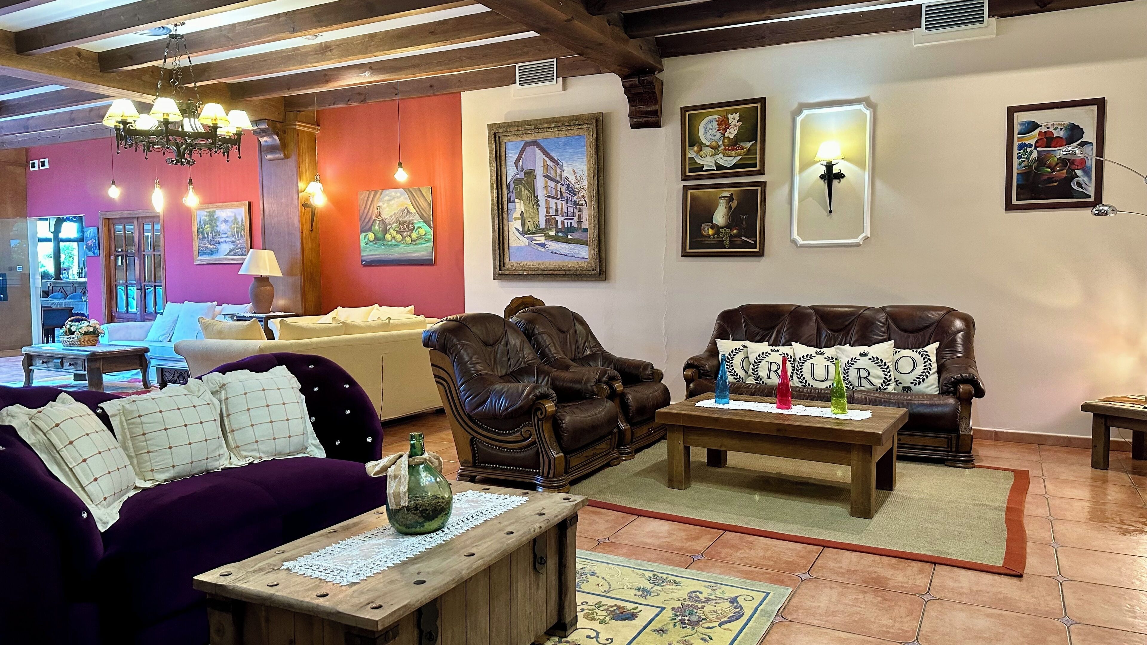 A cozy living room with a purple sofa, a brown leather armchair, and a wooden coffee table. There are paintings on the walls and a chandelier hanging from the ceiling.