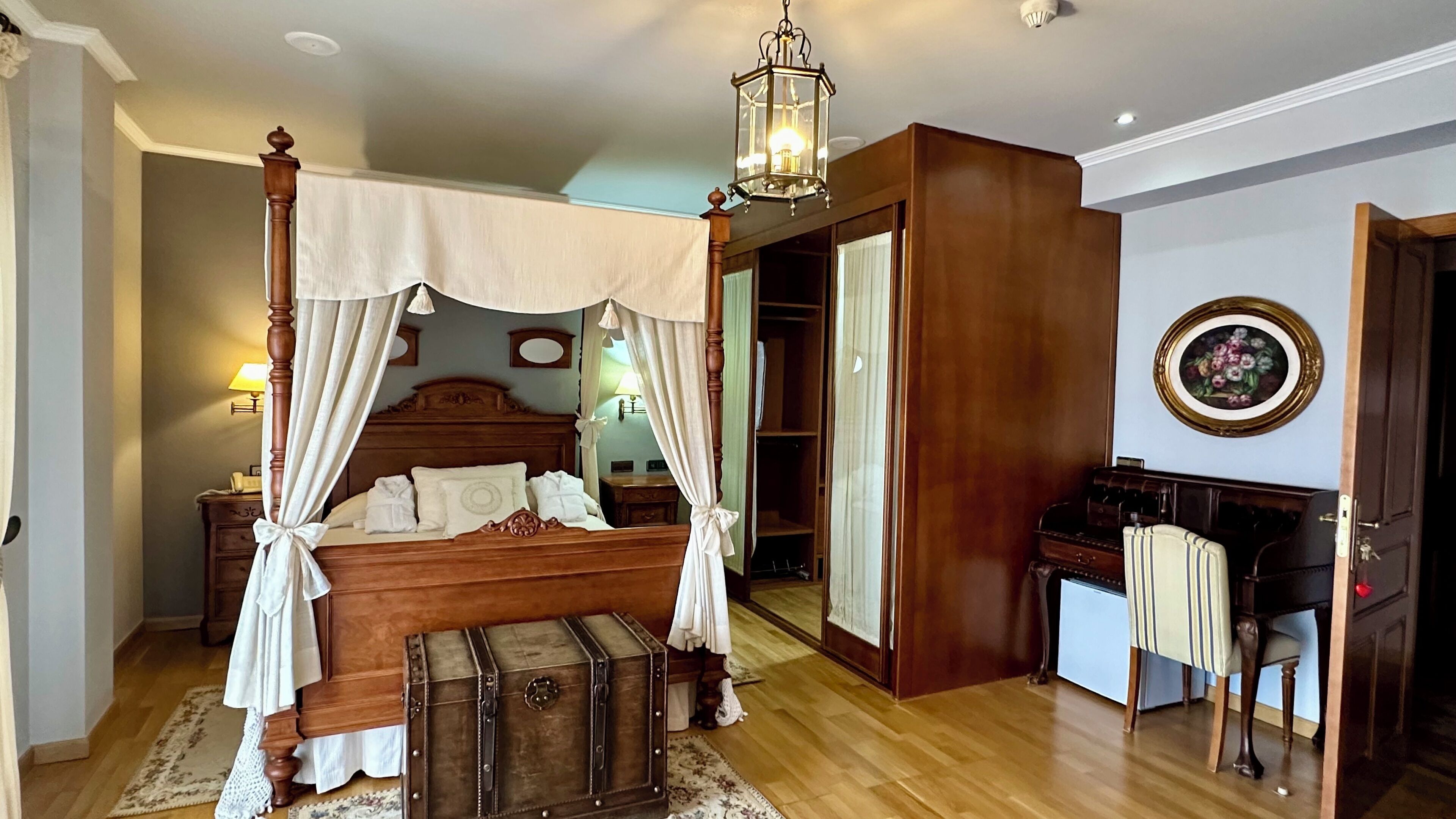 A bedroom with a four-poster bed, a canopy, a vintage suitcase, and a wooden desk with a chair.