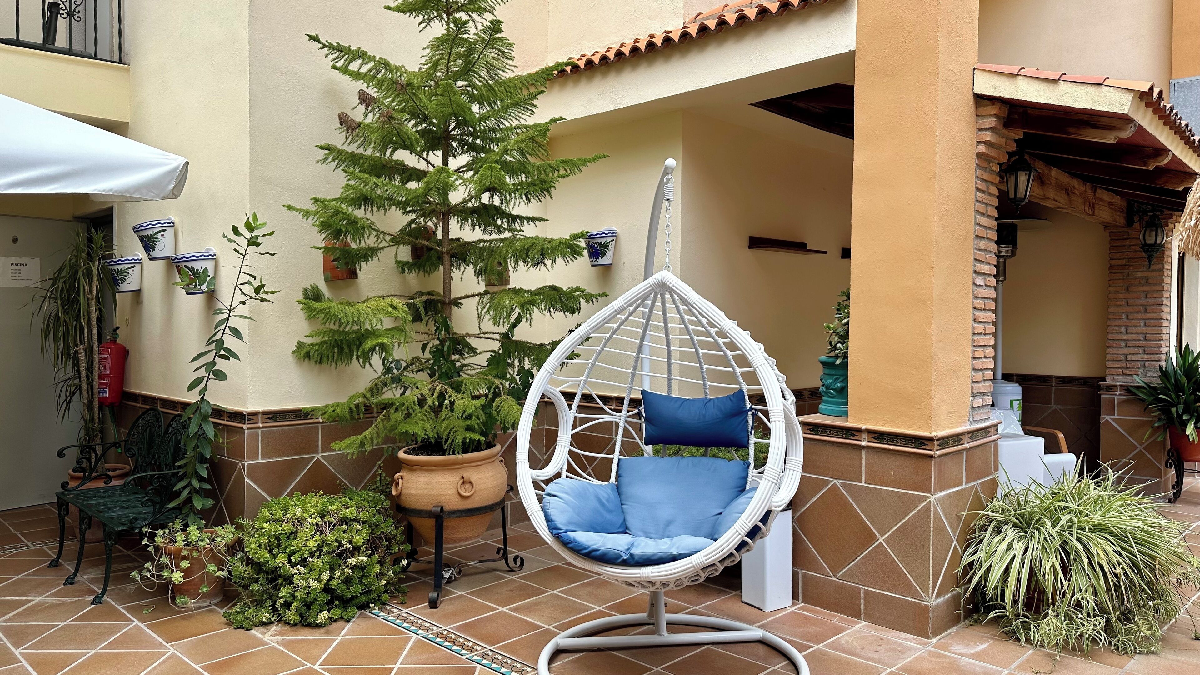 Una silla colgante con cojines azules en un patio con plantas en macetas y suelo de baldosas.