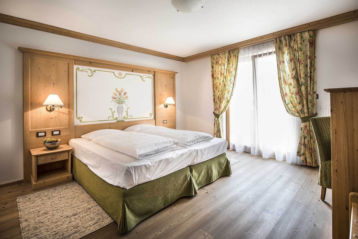 A bedroom with a bed, wooden headboard, two bedside lamps, a nightstand, a green skirted bed base, a floral curtain, and a wooden chair.