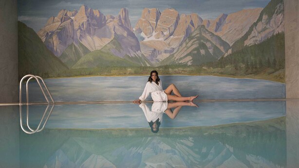 A person sitting on a pool edge with a mountainous landscape in the background. - Hotel Cristallo (Dobbiaco)