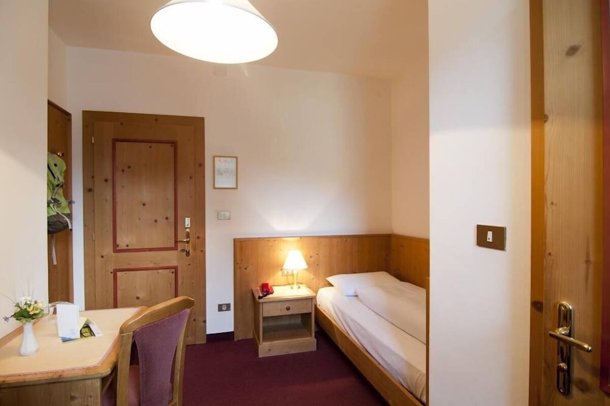 A small, cozy room with a wooden door, a bedside table with a lamp, a single bed, and a chair.