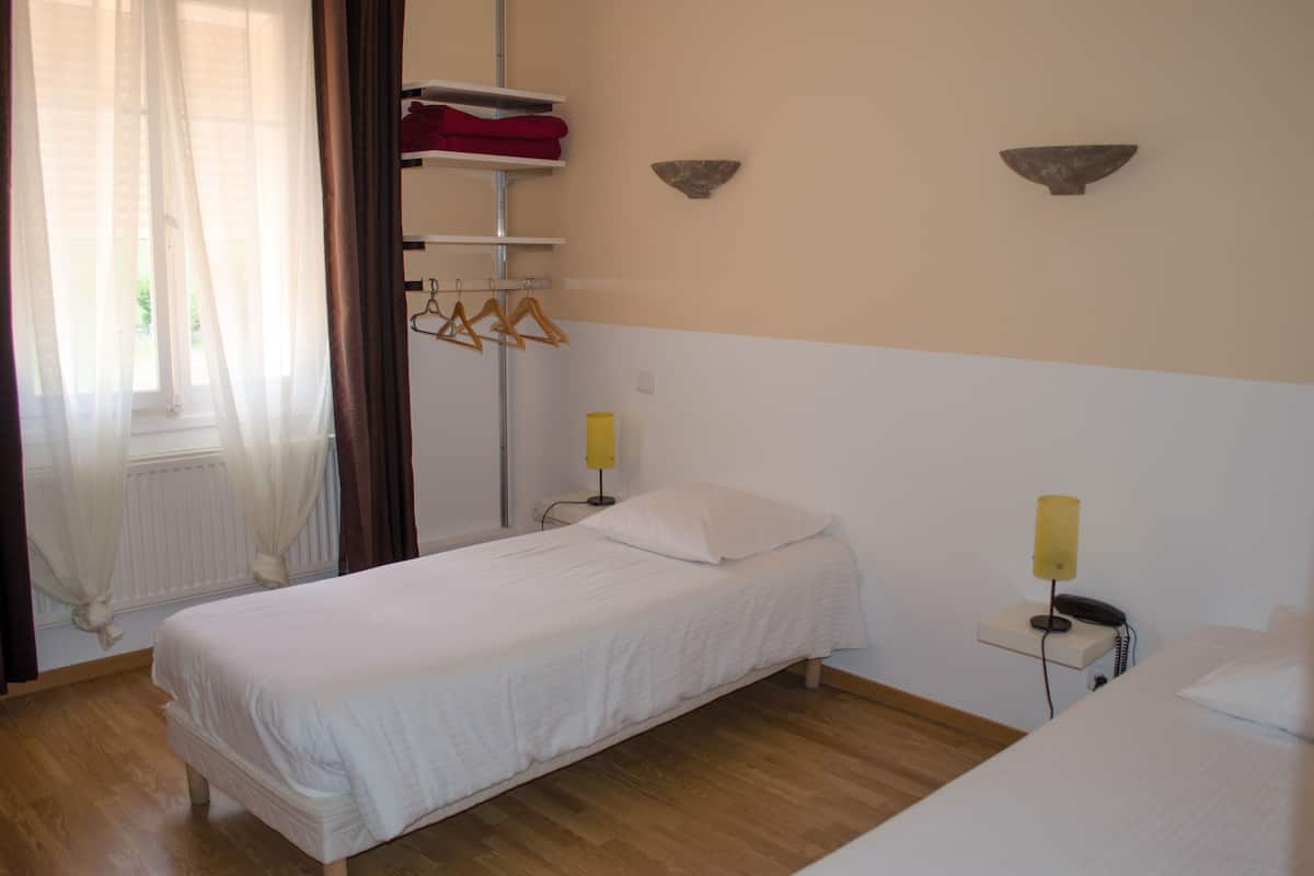 Een enkel bed met een witte matras en kussens, een nachtkastje met een lamp, een plank met rode handdoeken en een raam met dunne gordijnen.