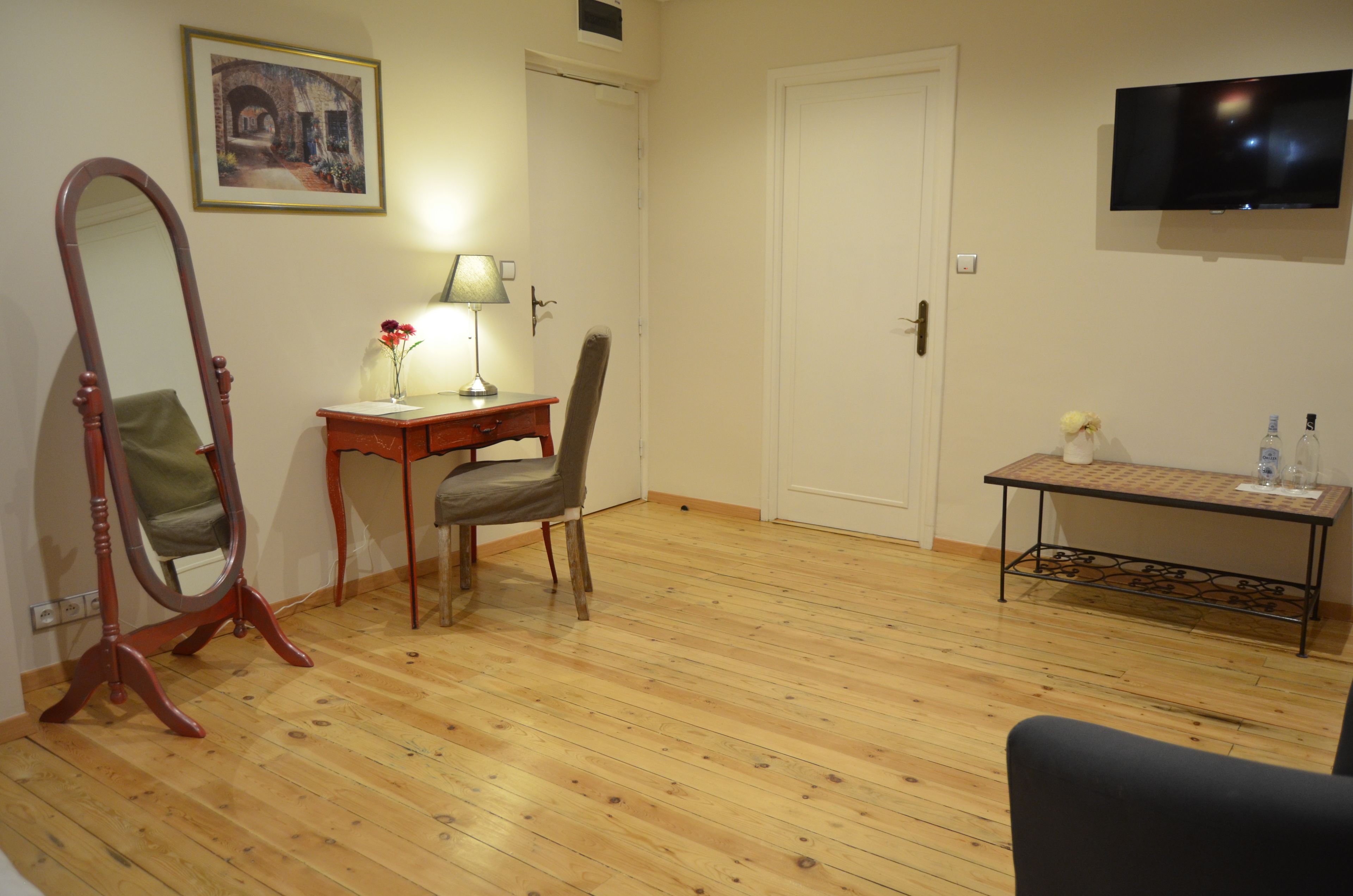 A room with wooden flooring, a desk with a lamp, a chair, a mirror, a television, and a small table with bottles.