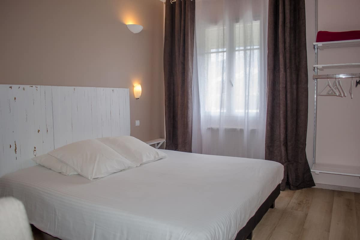 Een slaapkamer met een bed, een wit hoofdeinde, een wandlamp, een raam met gordijnen en een plank met een rood object.