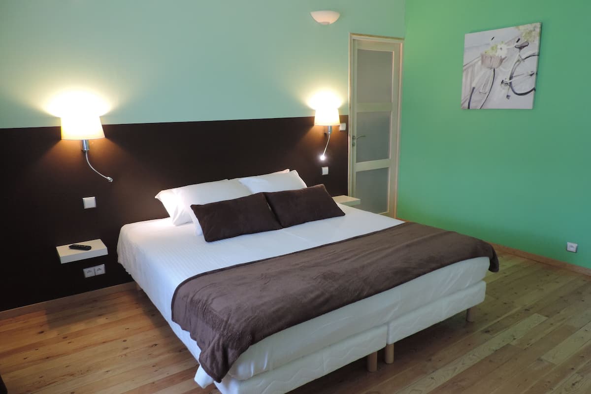 Een net opgemaakt bed met wit en bruin beddengoed, twee bedlampen, een nachtkastje en een groene muur met een schilderij.