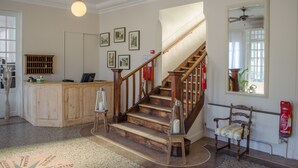Een trap met houten leuningen, een receptiebalie, ingelijste foto's aan de muur en een brandblusser in de buurt van een raam.