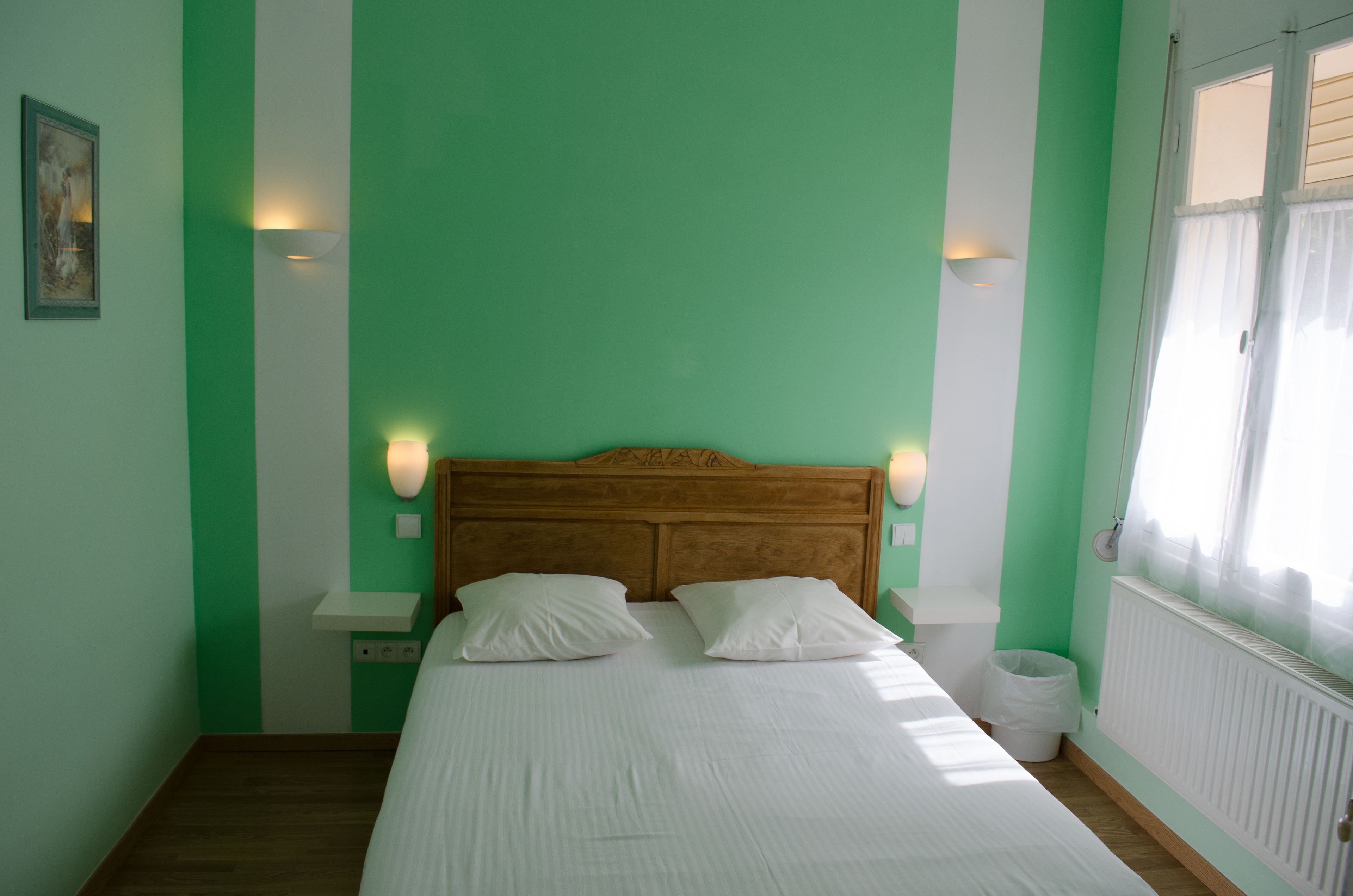 A bedroom with a bed, two bedside tables, a nightstand, a wall-mounted light, a window with curtains, and a radiator.