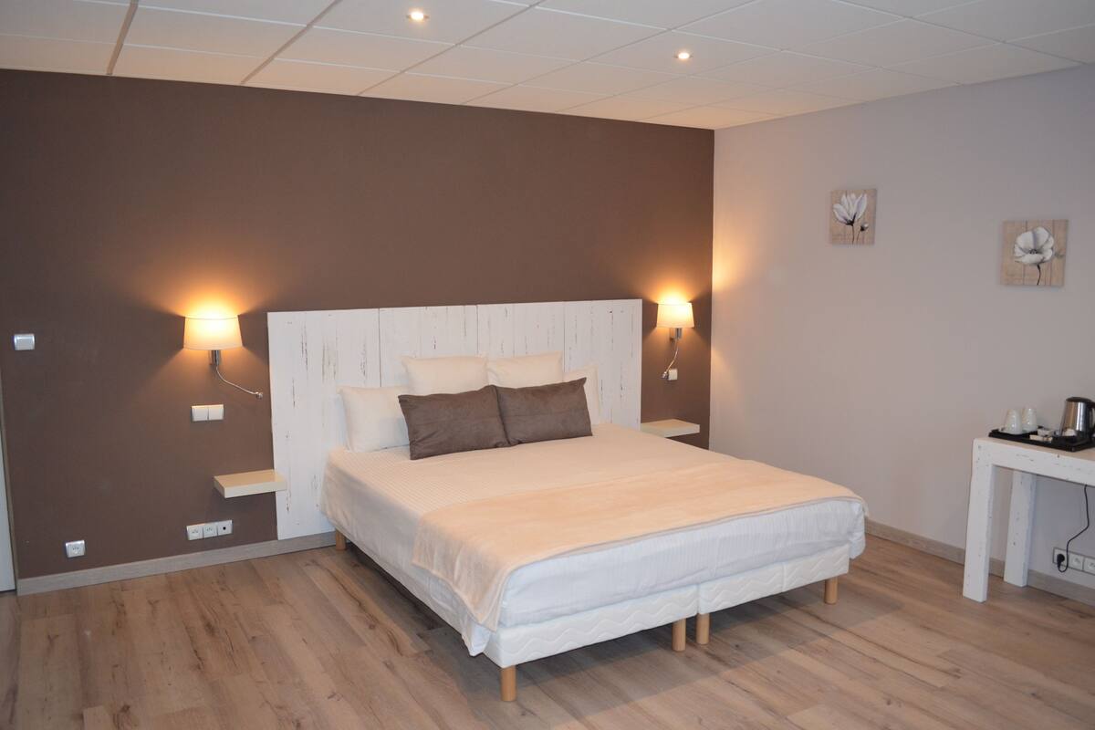 Een slaapkamer met een groot bed, twee bedlampen, een klein tafeltje met een waterkoker en een muur met twee bloemschilderijen.