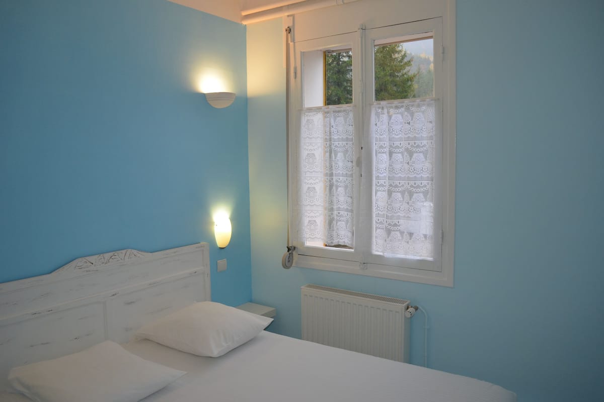 Een slaapkamer met een bed, witte kussens, een nachtkastje, een wandlamp en een raam met kantgordijnen.