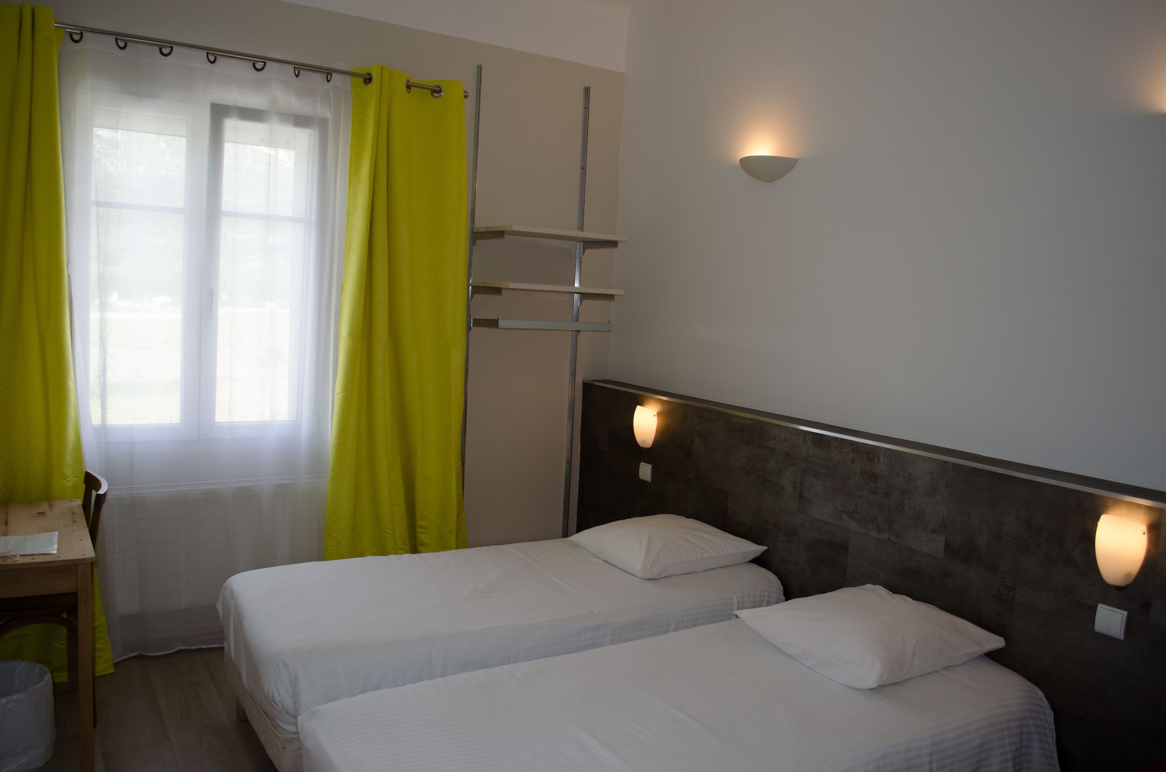 A room with two beds, a desk, and a window with yellow curtains.