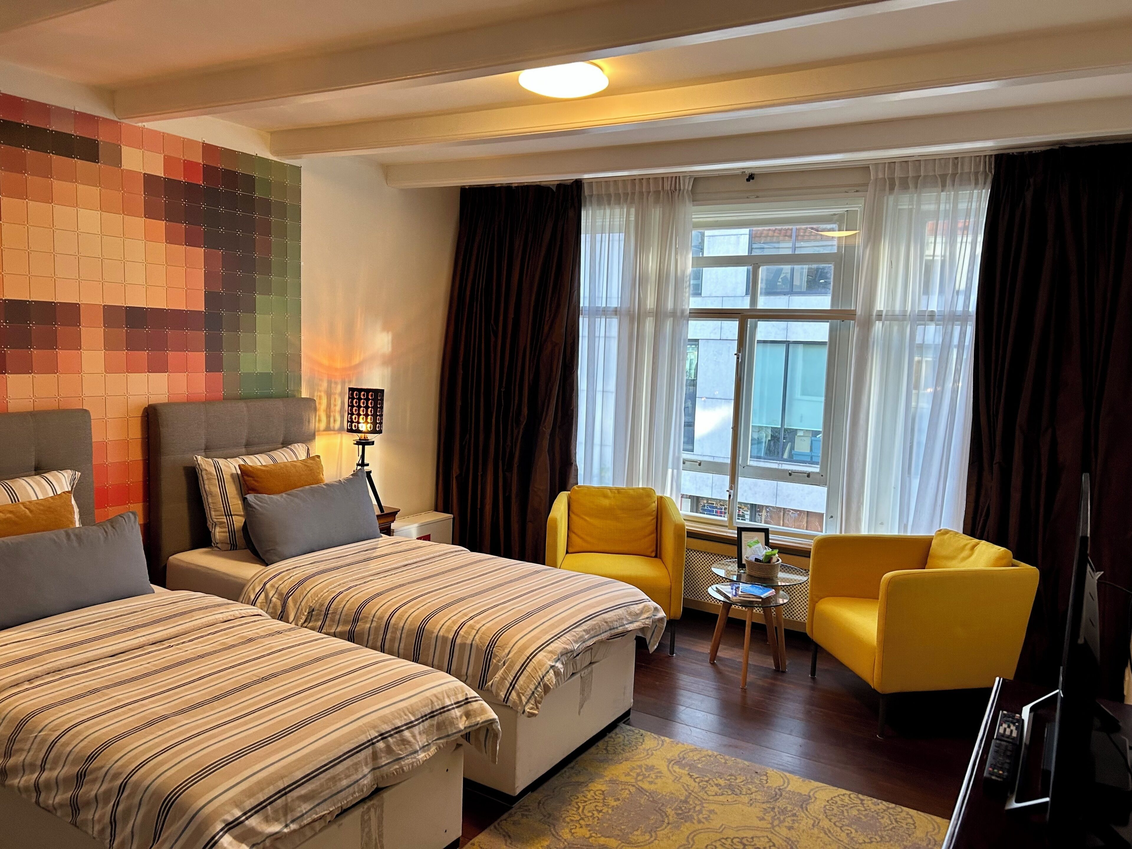 A hotel room with two beds, a yellow armchair, a small table, and a window with curtains.