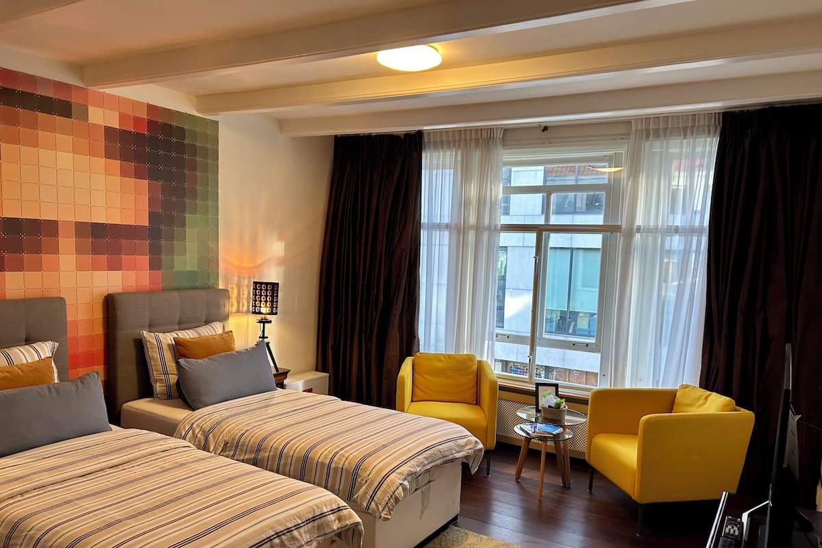 Habitación de hotel con dos camas, un sillón amarillo, una mesita y una ventana con cortinas.