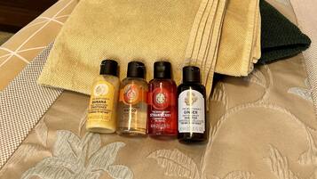 Een bed met een beige en witte bedspreide, een opgevouwen beige handdoek en vier flesjes The Body Shop producten op het bed.