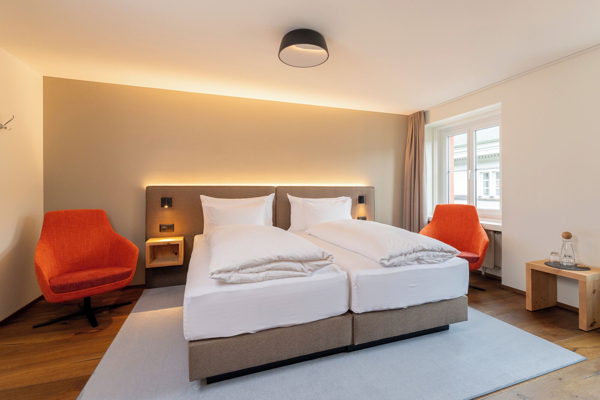 Habitación de hotel con una cama doble, dos mesitas de noche, dos sillas, una mesita pequeña y una ventana.