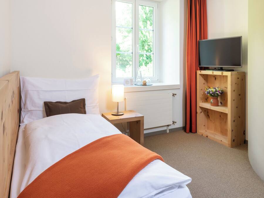 En pænt redt seng med hvidt og orange sengetøj, et natbord med lampe, et fjernsyn på et træstel og et vindue med gardiner.