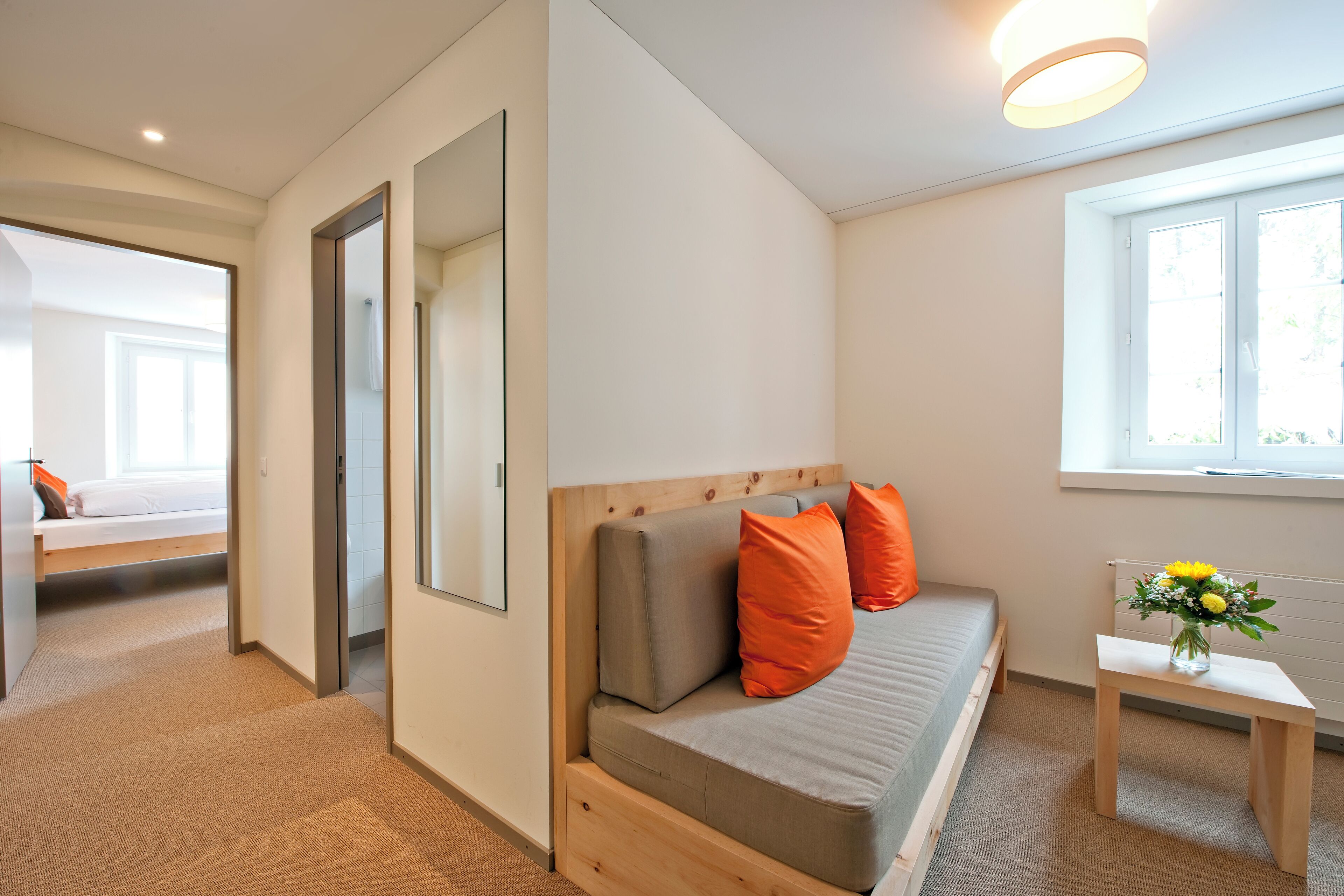 A modern bedroom with a wooden headboard, a grey mattress, and orange pillows. There is a small wooden table with a vase of flowers next to a window.