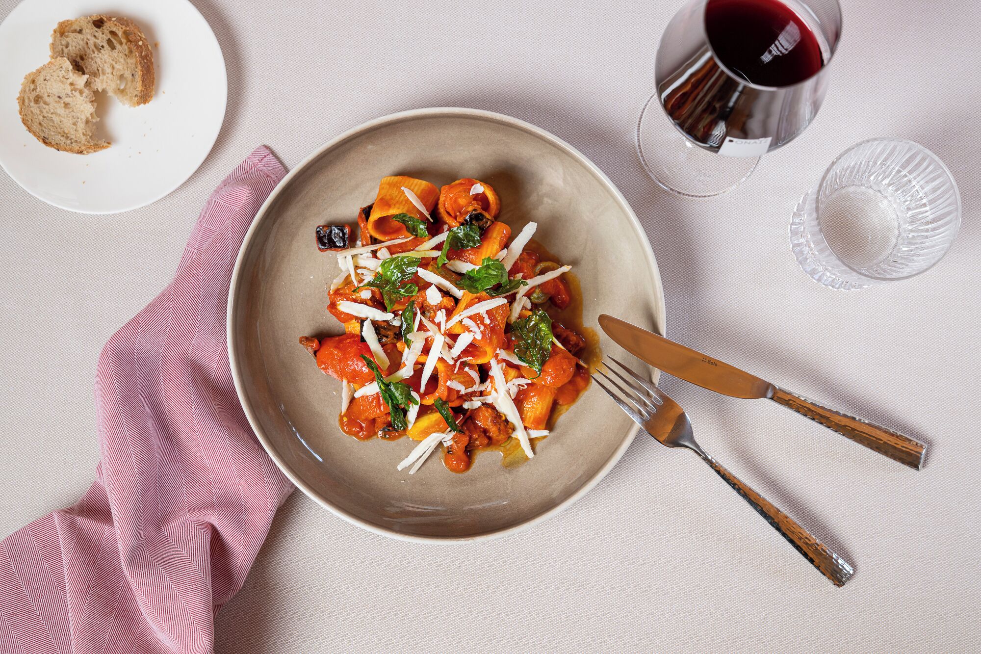 Un plato de pasta con queso y vegetales verdes, una copa de vino tinto y un tenedor sobre una mesa con servilleta.