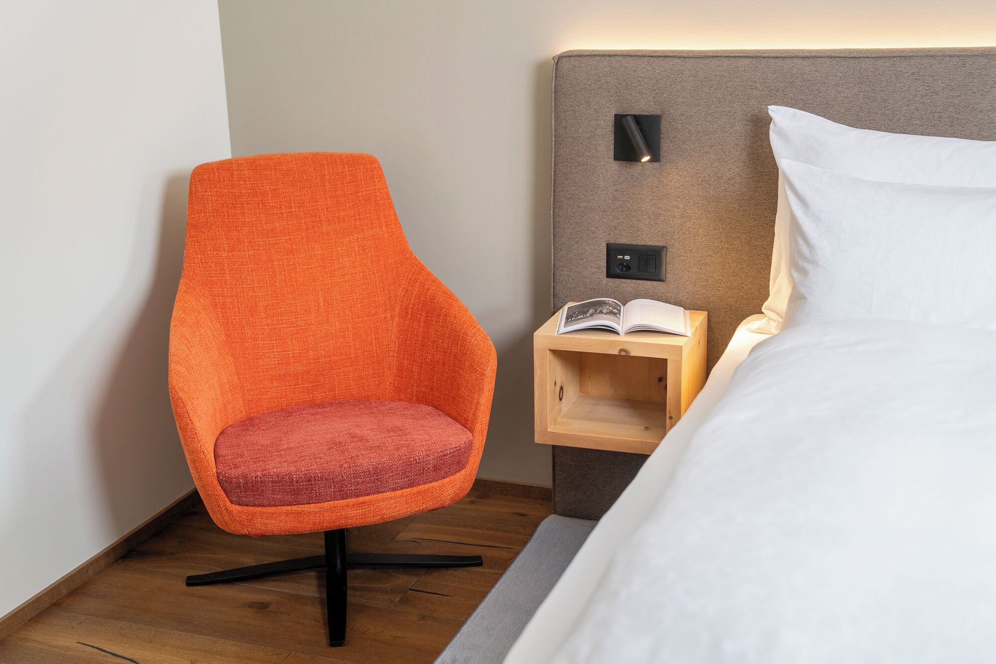 Una camera d'albergo con una poltrona arancione, un comodino in legno con un libro e un letto con lenzuola bianche.