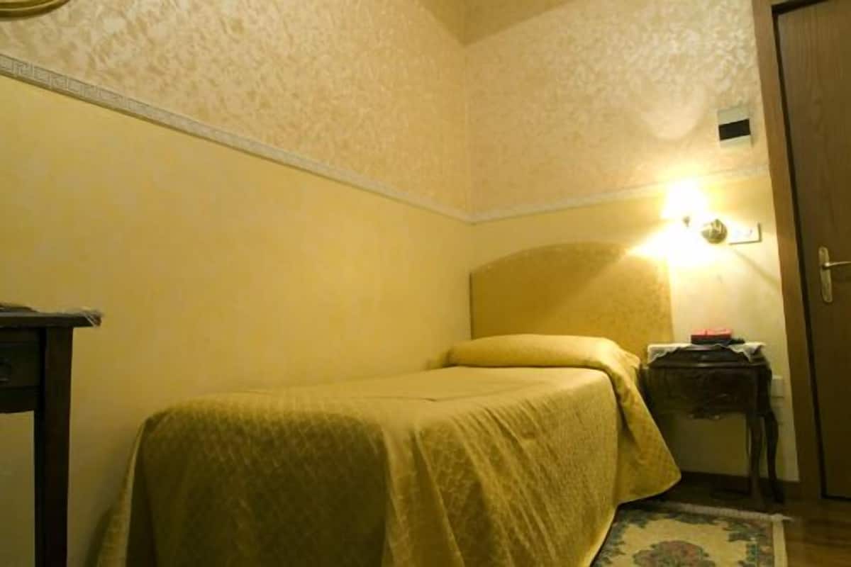 A small, single-bed room with a yellow bedspread, a bedside table with a lamp, and a door to the right.