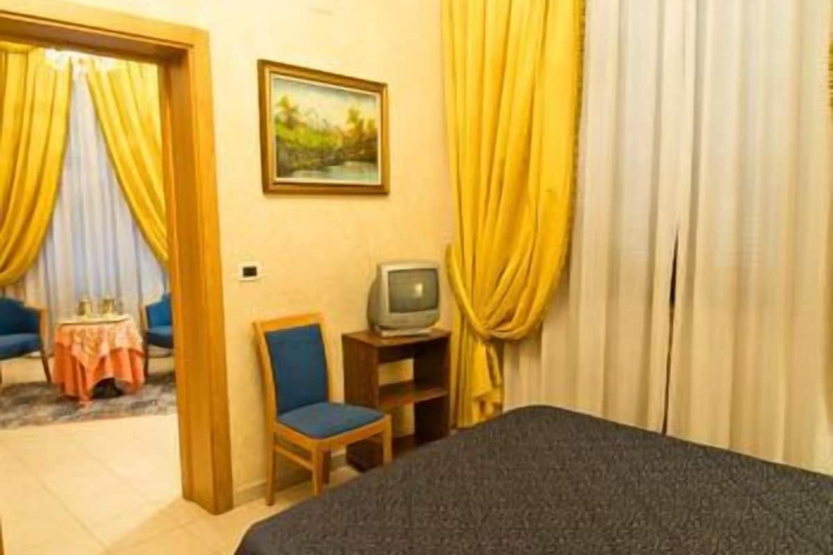 A hotel room with a bed, a television on a stand, a small table with a chair, and a painting on the wall.