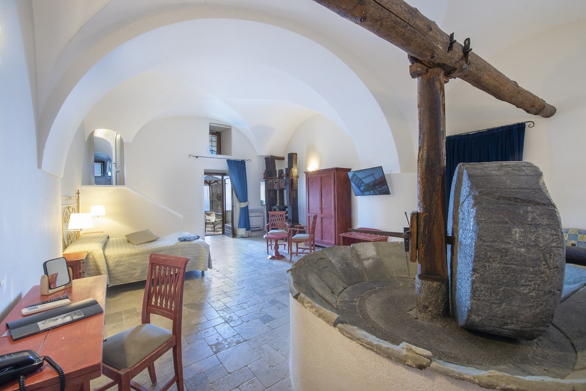 Una habitación con un pozo de piedra, vigas de madera, una cama, un escritorio con lámpara y un televisor.
