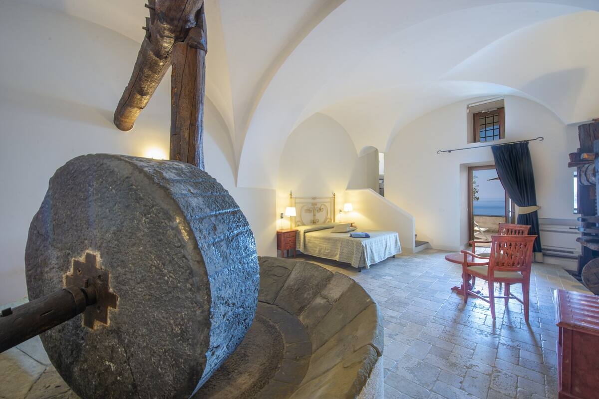 Un molino de piedra, una cama con cabecera, una silla y una ventana con cortinas.